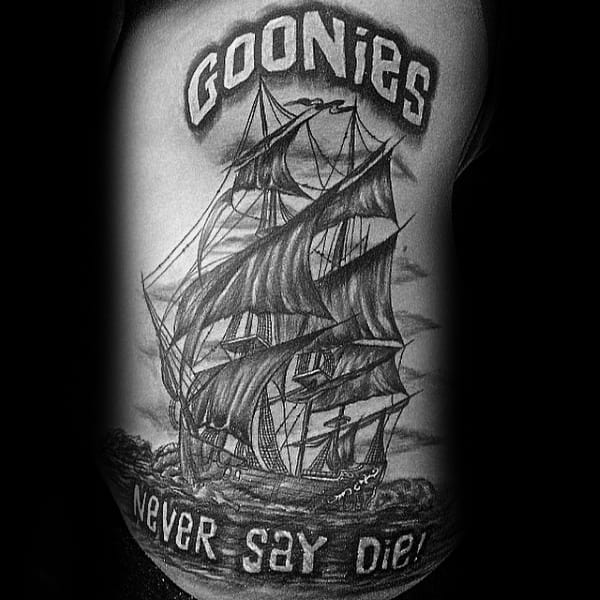 Black and gray tattoo of a detailed pirate ship from The Goonies, with bold lettering reading "Goonies Never Say Die!".