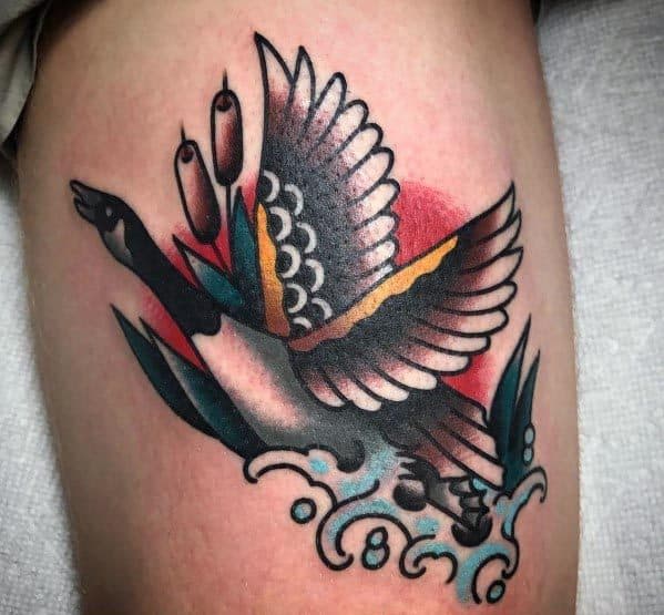 Traditional-style tattoo of a flying goose with cattails, water splashes, and a red sunburst background, featuring bold black outlines and vibrant colors.