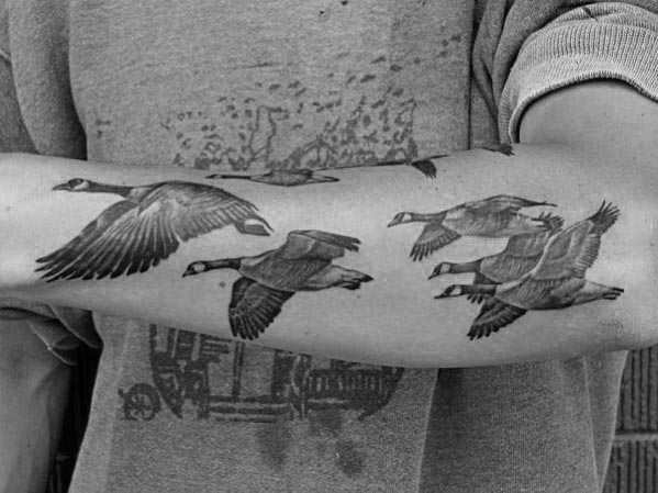 Black and gray tattoo of a flock of geese in flight, inked in a realistic style on the forearm, with detailed feathers and shading.