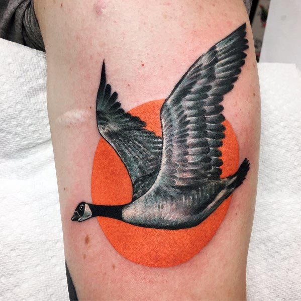 Realistic tattoo of a flying goose with detailed black and gray shading, set against a bold orange circle background, inked on the upper arm.