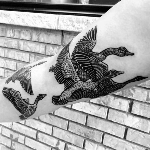 Black and gray tattoo of a flock of geese in flight, inked on the forearm with intricate linework and detailed feather shading.