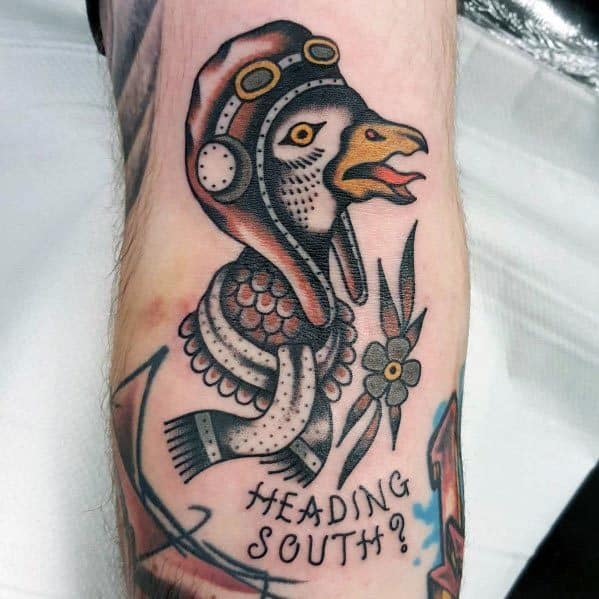 Traditional-style tattoo of a goose wearing an aviator helmet and scarf, with the phrase "HEADING SOUTH?" below, accented with a flower and arrow.