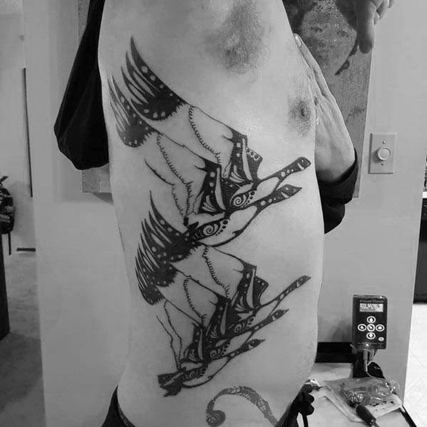 Black and gray tattoo of three intricately designed geese in flight, inked on the ribcage with bold patterns and geometric details.
