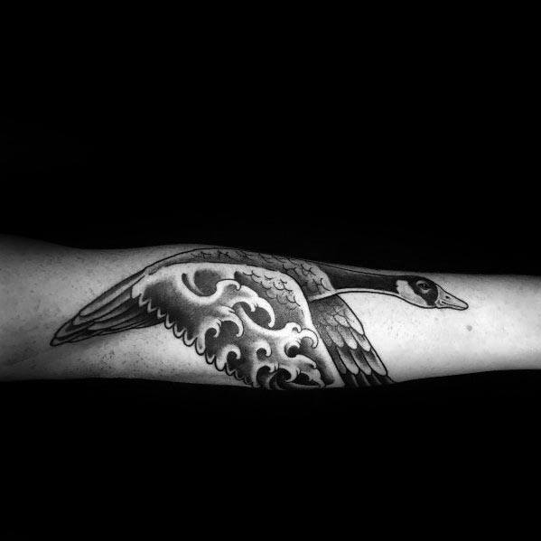 Black and gray tattoo of a flying goose with intricate shading and stylized cloud-like patterns on its wings, inked on the forearm.