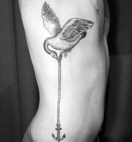 Black and gray tattoo of a goose in flight gripping a rope with its beak, connected to an anchor, inked on the ribcage with fine shading.