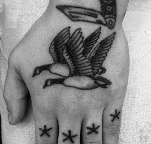 Black and gray tattoo of two geese flying in unison, inked on the hand with bold lines and shading, complemented by small star symbols on the fingers.
