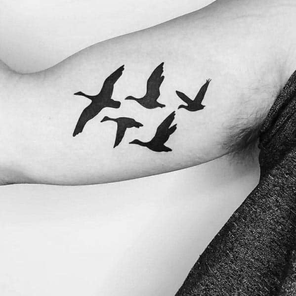Minimalist black silhouette tattoo of a flock of geese in flight, inked on the inner arm, creating a simple and elegant design.