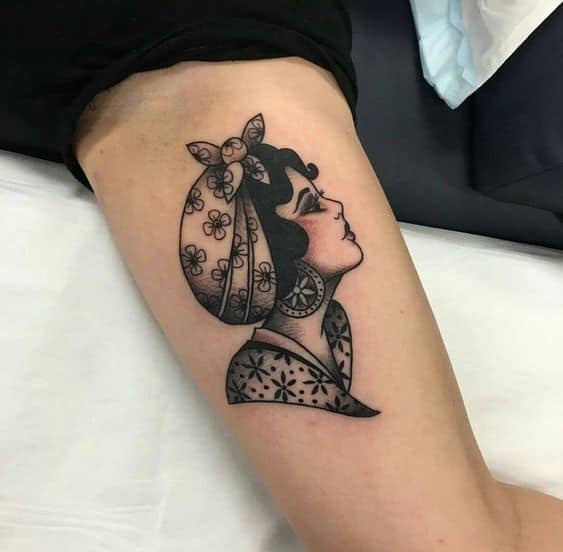 Black and grey tattoo of a gypsy woman in profile with a floral headscarf, large earrings, and delicate shading on the upper arm.