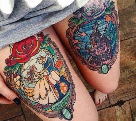 Stained glass-style thigh tattoos featuring Belle and the Beast dancing in one frame and the Beast’s castle in the other, with intricate details and bold colors.