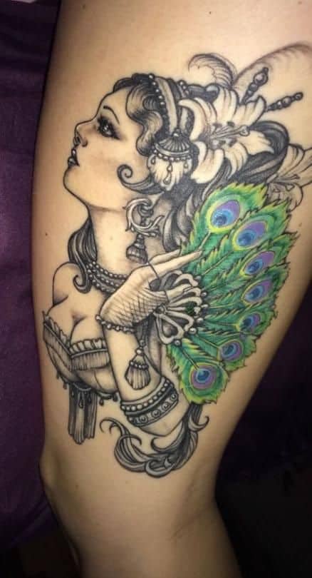 Ornate thigh tattoo of a woman with intricate jewelry, feathered accessories, and a vibrant peacock feather fan, blending black and grey shading with rich green and blue hues.
