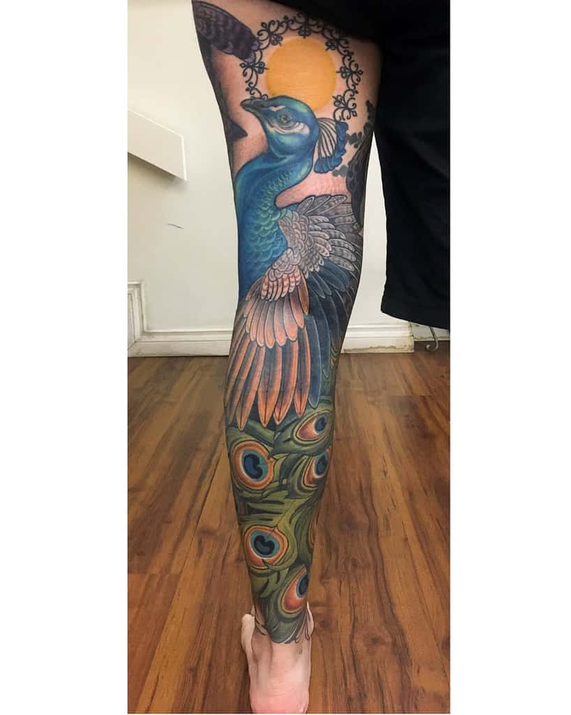 Full-sleeve peacock tattoo with vibrant colors, featuring a detailed bird and a peacock tail adorned with rich blue and green hues, set against a sun-like backdrop.