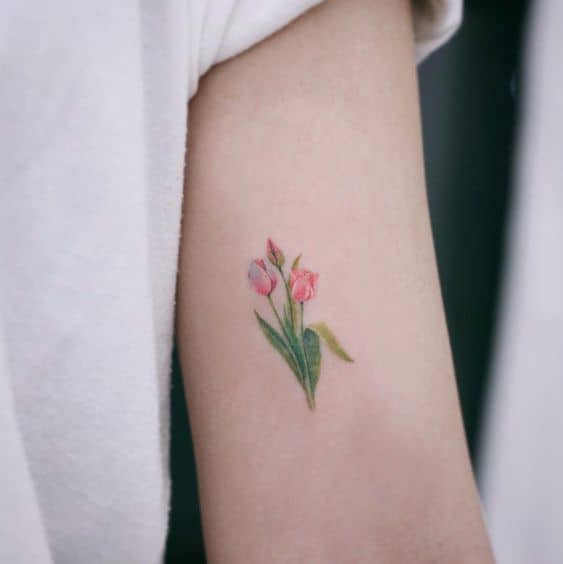 A delicate watercolor tulip tattoo on the upper arm, featuring three small pink flowers with soft green leaves for a natural and elegant look.