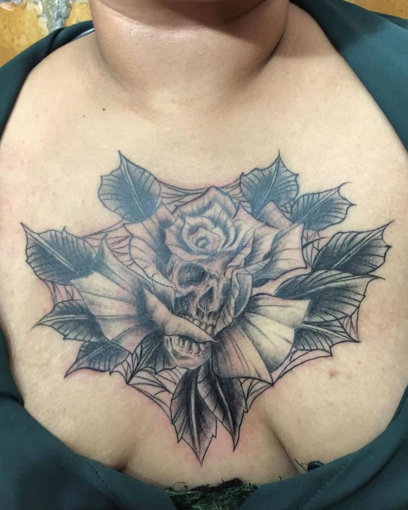 Black and grey chest tattoo featuring a skull within a blooming rose, surrounded by leaves and a subtle spiderweb design.