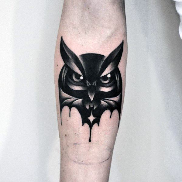 Bold black and grey gradient owl tattoo with sharp eyes and stylized wings forming a dripping effect on the forearm.