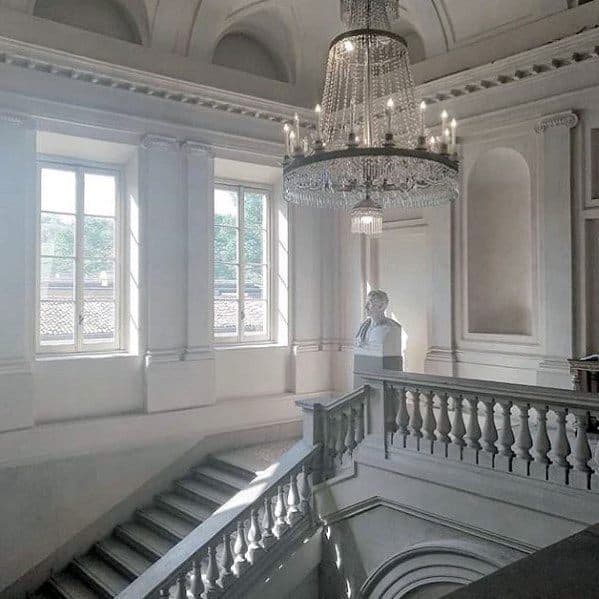 A grand staircase with elegant crown molding, a stunning chandelier, and classical architectural details, bathed in natural light