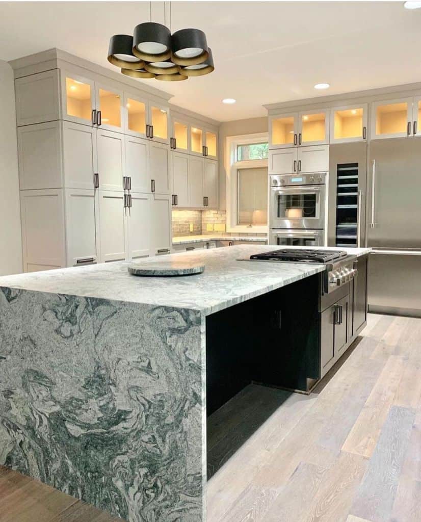 Elegant kitchen with gray cabinetry, illuminated glass panels, and a striking granite waterfall countertop.