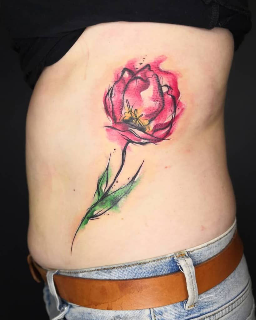 A vibrant watercolor tulip tattoo on the side of the torso, featuring bold pink petals, green leaves, and artistic splashes for a dynamic look.
