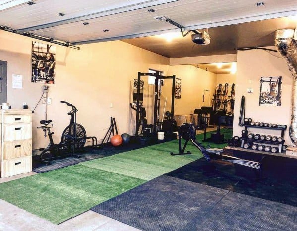 A garage gym with rubber flooring, turf, a rowing machine, air bike, squat rack, and dumbbell rack.