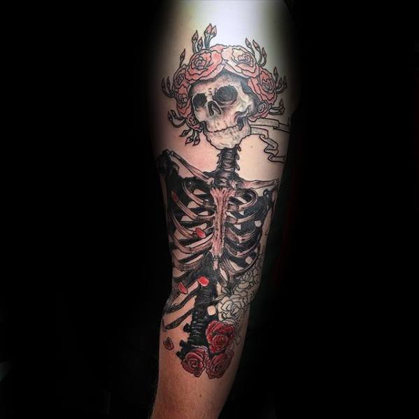 Grateful Dead skeleton tattoo with a crown of red roses and detailed ribcage, blending black and red ink on the upper arm.