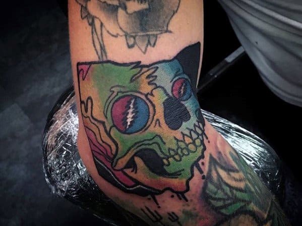 Colorful Grateful Dead skull tattoo with bold outlines, featuring lightning bolt eyes and a melting rainbow effect on the elbow.