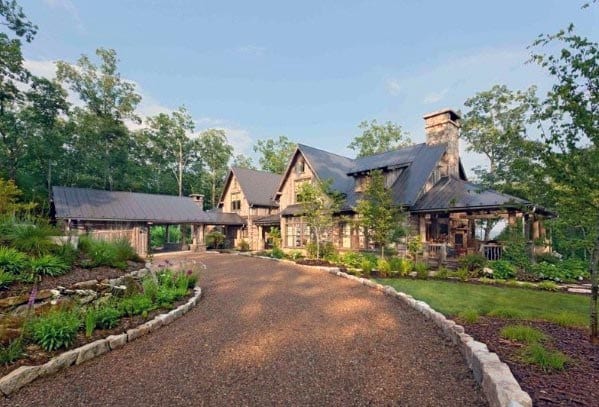 Gravel driveway with natural stone edging leading to a rustic house surrounded by greenery.