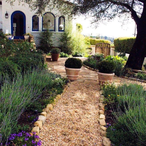 A charming gravel pathway leading to a house, bordered by smooth stones, with lush plants and decorative pots along the sides