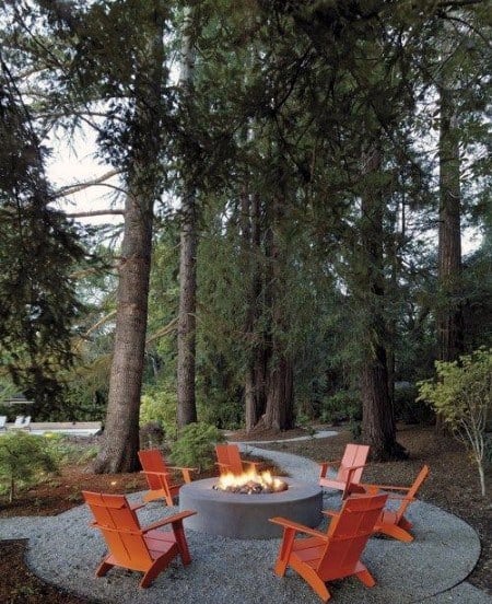 Red chairs encircle a cozy fire pit on gravel, nestled among tall trees