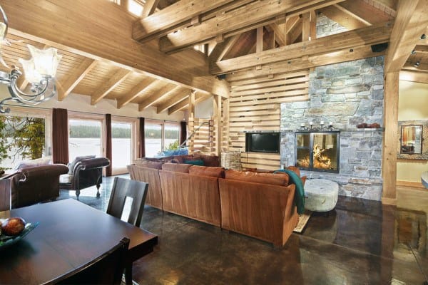 Spacious cabin interior with wooden beams, stone fireplace, large windows, and a sleek concrete floor overlooking a lake