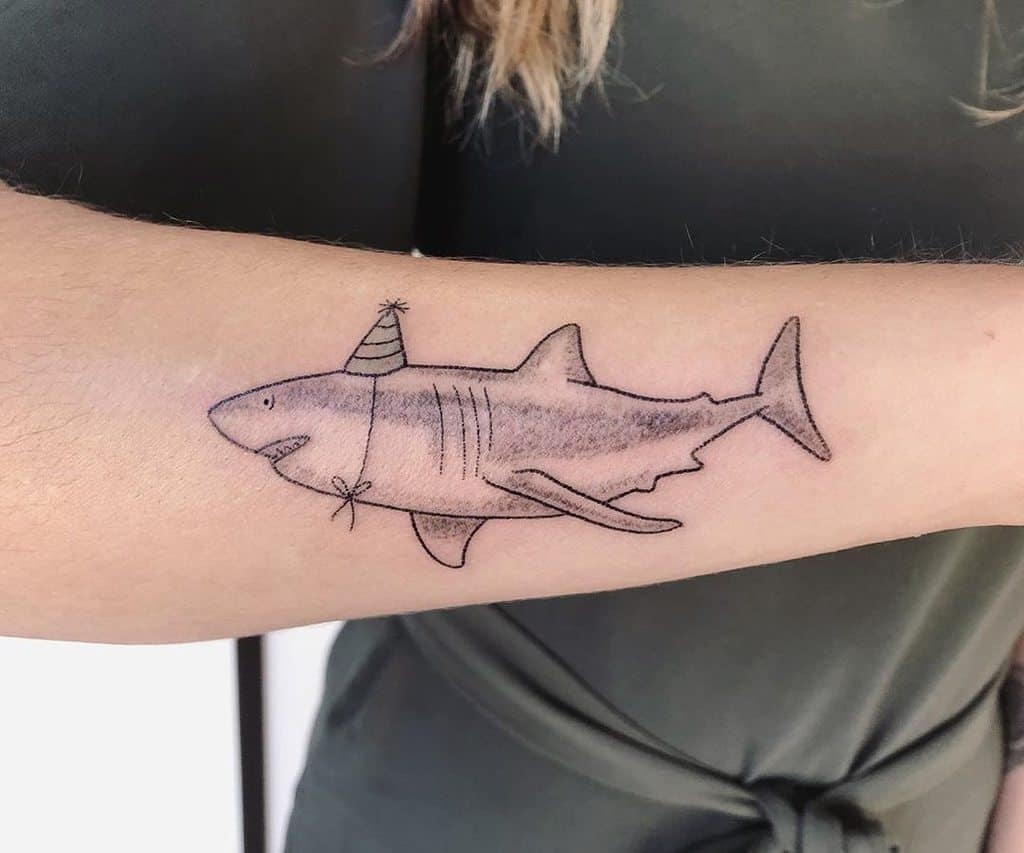 A black ink tattoo of a shark wearing a small party hat, drawn in a simple and playful style on the forearm.
