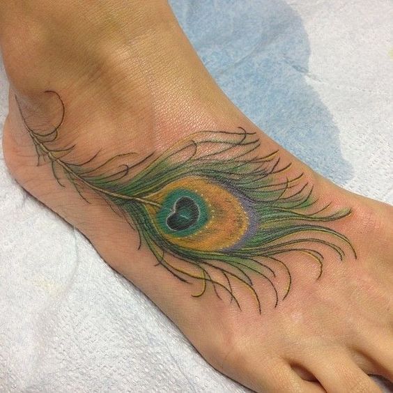 Vibrant peacock feather tattoo on the foot, featuring rich green, blue, and gold hues with intricate shading and fine line details.