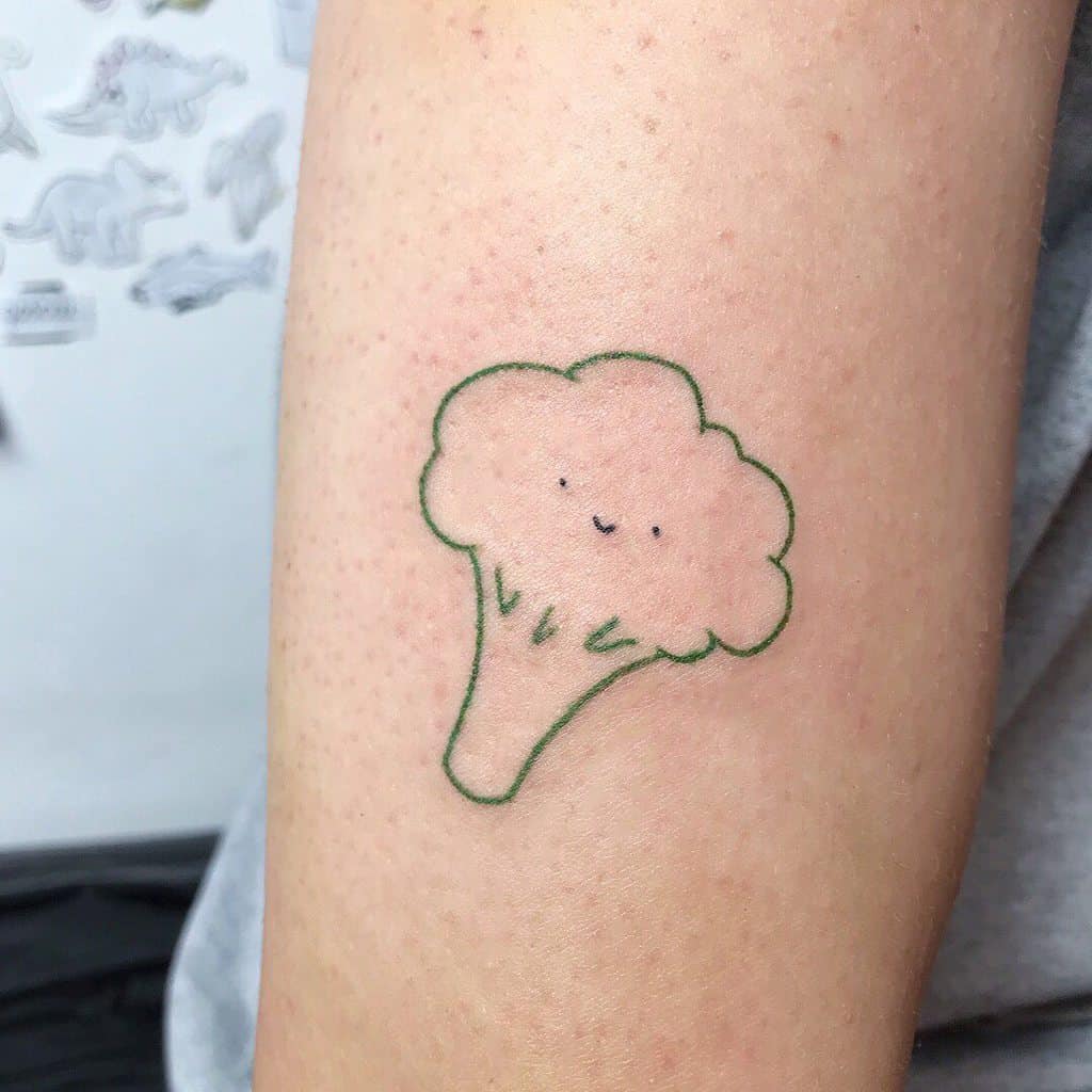 A minimalist tattoo of a smiling broccoli with a simple green outline and tiny facial features, creating a cute and playful design.
