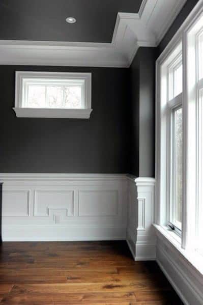 A stylish dining room with dark grey walls, white crown molding, and wainscoting, complemented by natural wood floors and large windows
