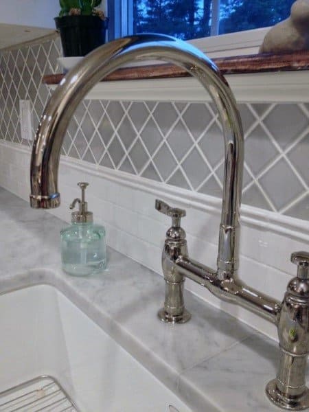 Silver kitchen faucet with marble countertop and diamond-patterned white tile backsplash.