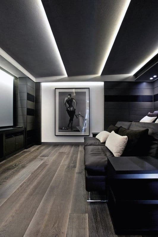 Modern living room with dark walls, wooden floors, a large photo, home theater, and a black sofa with white pillows
