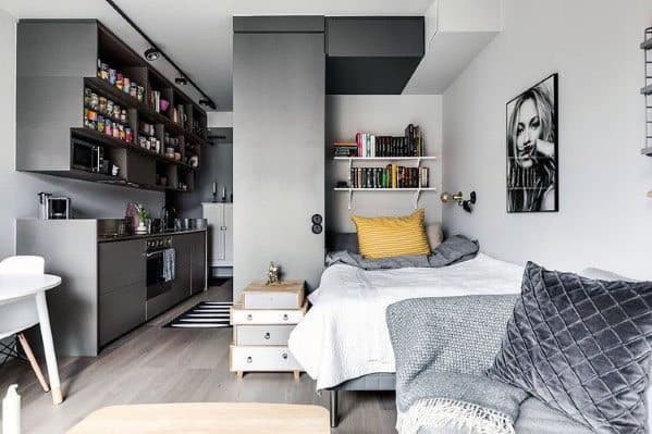 Modern studio with gray kitchen, cozy bed, open shelving, and hardwood floors.