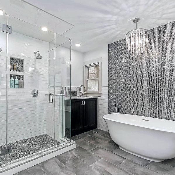 Elegant grey bathroom featuring a freestanding tub, mosaic accent wall, glass-enclosed shower, and a crystal chandelier.