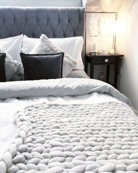A cozy bedroom with grey plush pillows, a chunky knit blanket, and a lit bedside table lamp