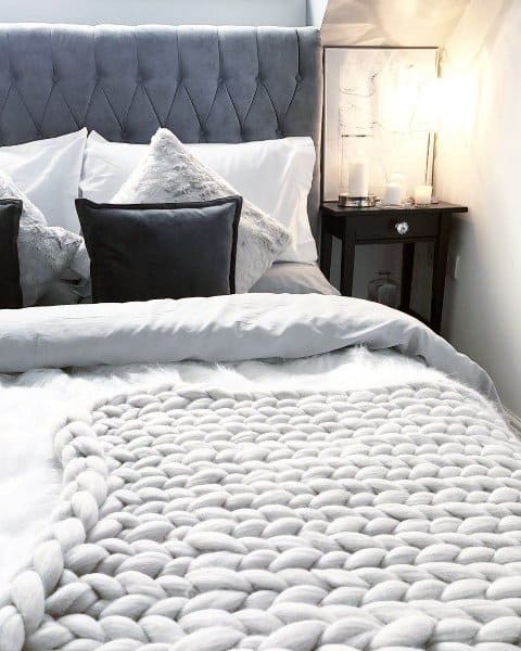 Cozy bedroom with a plush grey headboard, pillows, a chunky knit blanket, and a lit bedside lamp