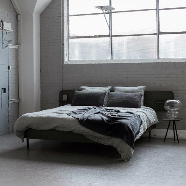 Minimalist bedroom with a low bed, grey bedding, and a side table by large industrial-style windows