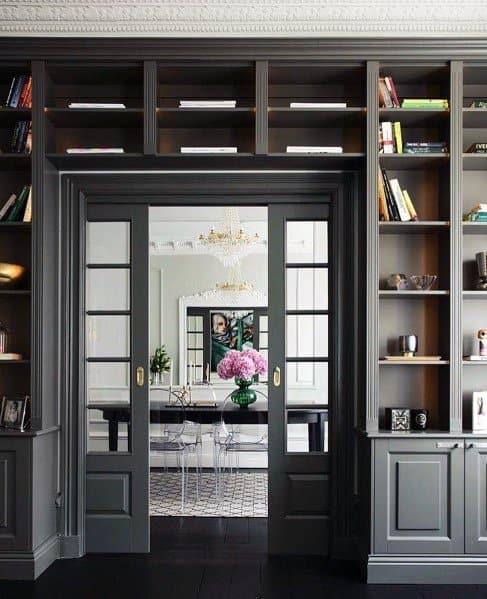 Elegant room with gray built-in bookcases framing a glass-paneled doorway, leading to a dining room with a chandelier