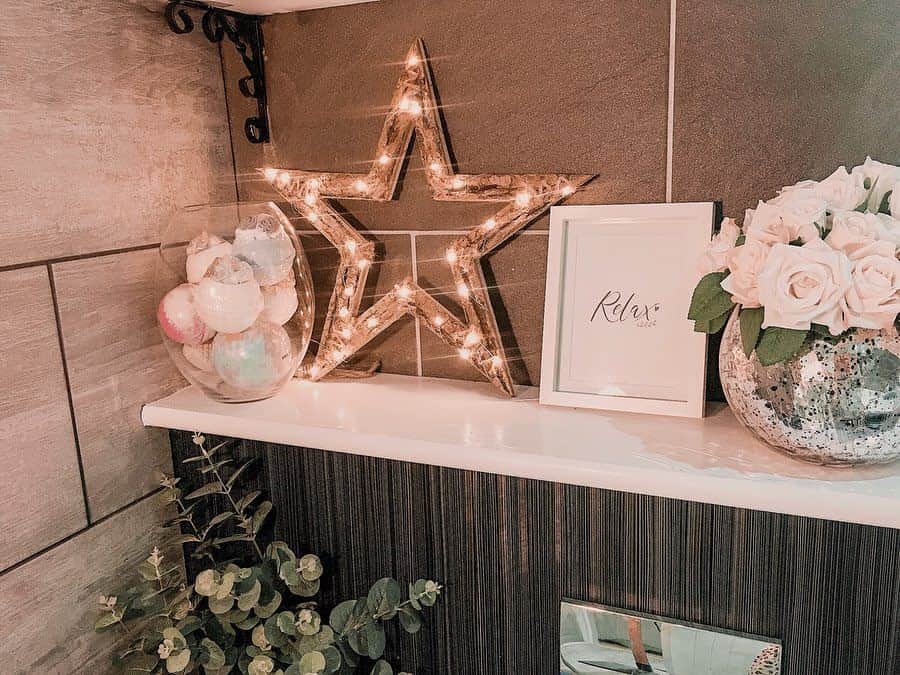 White bathroom shelf with a lit star, vase of roses, framed "Relax" sign, and glass jar of bath bombs.