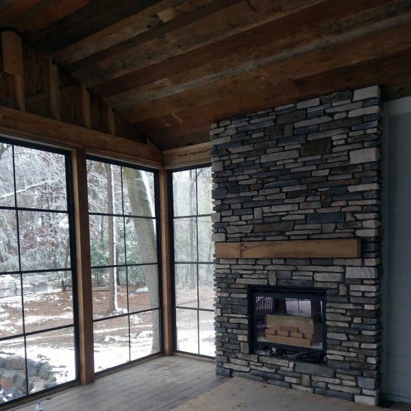 Rustic sunroom with grey stacked stone fireplace, wooden mantel, and large grid windows.