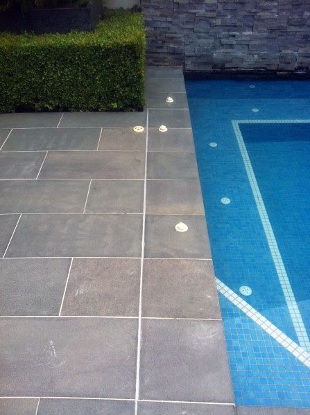 Blue mosaic pool tiles with sleek grey slate deck and trimmed hedge in a modern outdoor design.