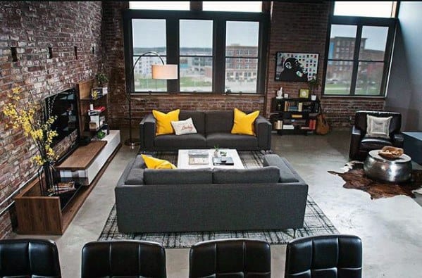 Dark grey upholstered sofas with yellow accent pillows in an industrial-style living room with exposed brick walls and large windows.
