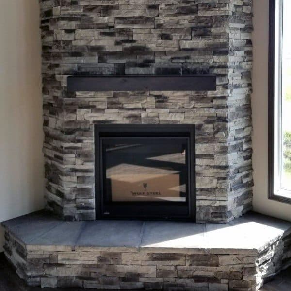Modern gray stacked-stone corner fireplace with black mantel and tiled hearth.