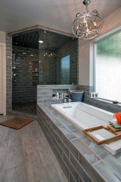 Modern bathroom with a sunken tub, corner glass-enclosed shower, grey tiles, wooden mat, and spherical ceiling light
