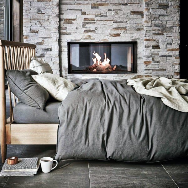 A cozy bedroom with a stone fireplace, wooden bed, and grey bedding; a cup and book rest on the grey floor