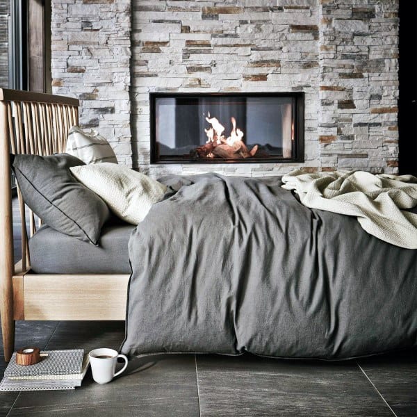 Cozy bedroom with grey bedding, a wooden bed, and a modern fireplace set in a stone wall