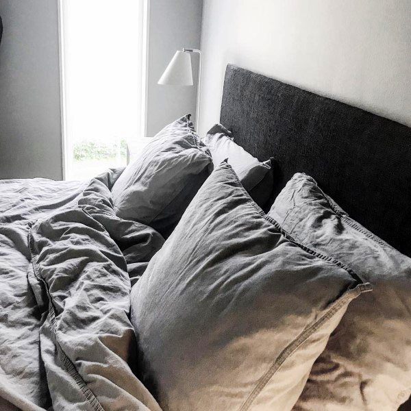 A cozy bed with grey pillows and blanket next to a tall window and wall-mounted lamp
