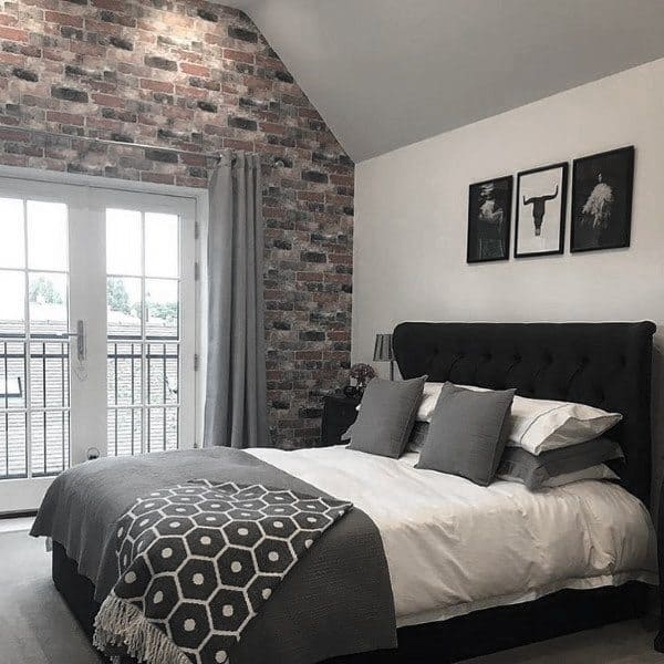 Cozy bedroom with a brick accent wall, a grey bed, and monochrome artwork; large windows fill the space with natural light